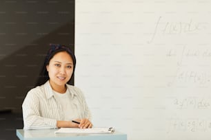 Portrait of content attractive Asian student girl standing at lecture stand with papers against whiteboard in classroom