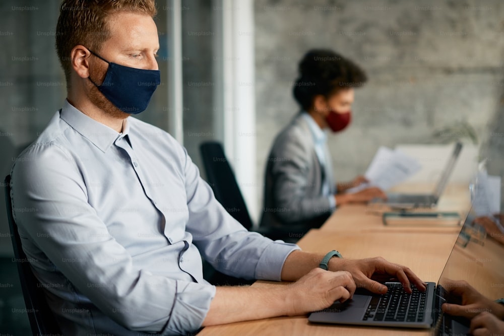 Uomo d'affari che indossa una maschera facciale mentre lavora su un computer in ufficio durante la pandemia di coronavirus.
