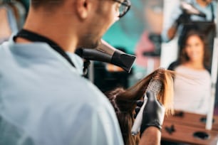 Primo piano del parrucchiere che usa l'asciugacapelli e la spazzola rotonda mentre acconcia i capelli del cliente in salone.