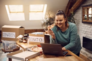 Young woman eating pizza and using laptop while collecting donations for charitable community.
