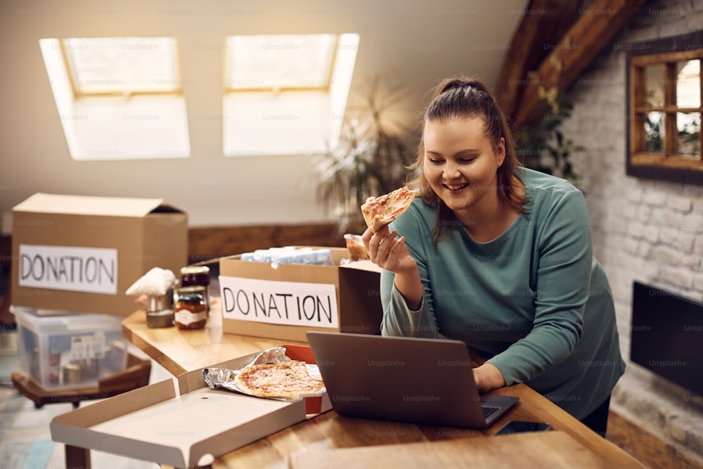 Jovem mulher comendo pizza e usando laptop enquanto coletava doações para a comunidade de caridade.