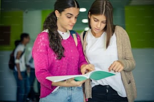 Two students girls reading book together.  Students standing in school hallway. Focus is on foreground.