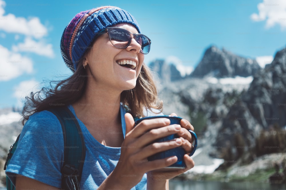 Portrait of beautiful hiking woman standing in stunning mountain wilderness with cup near snowy lake