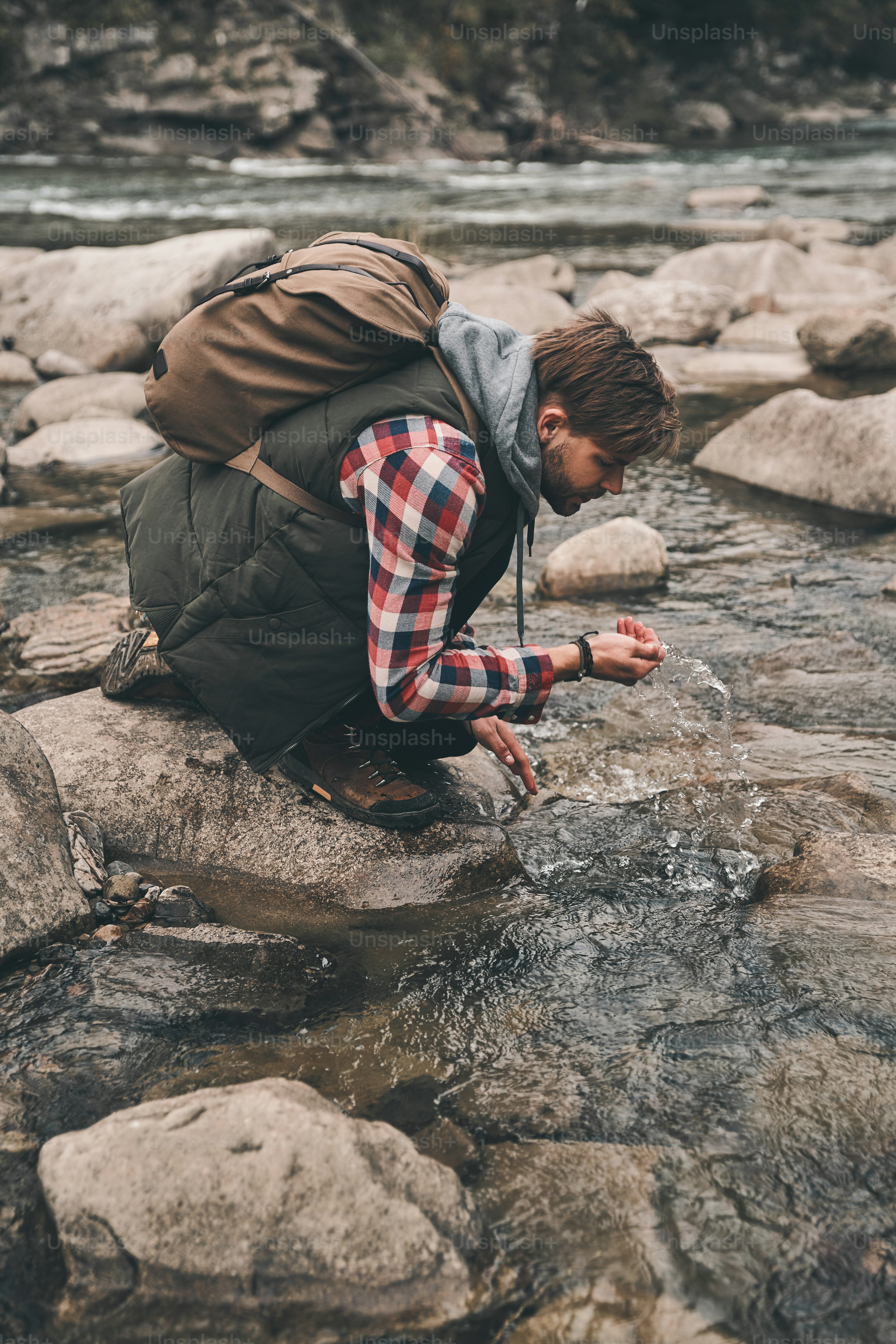 Beau jeune homme moderne buvant l’eau de la rivière lors d’une randonnée dans les montagnes