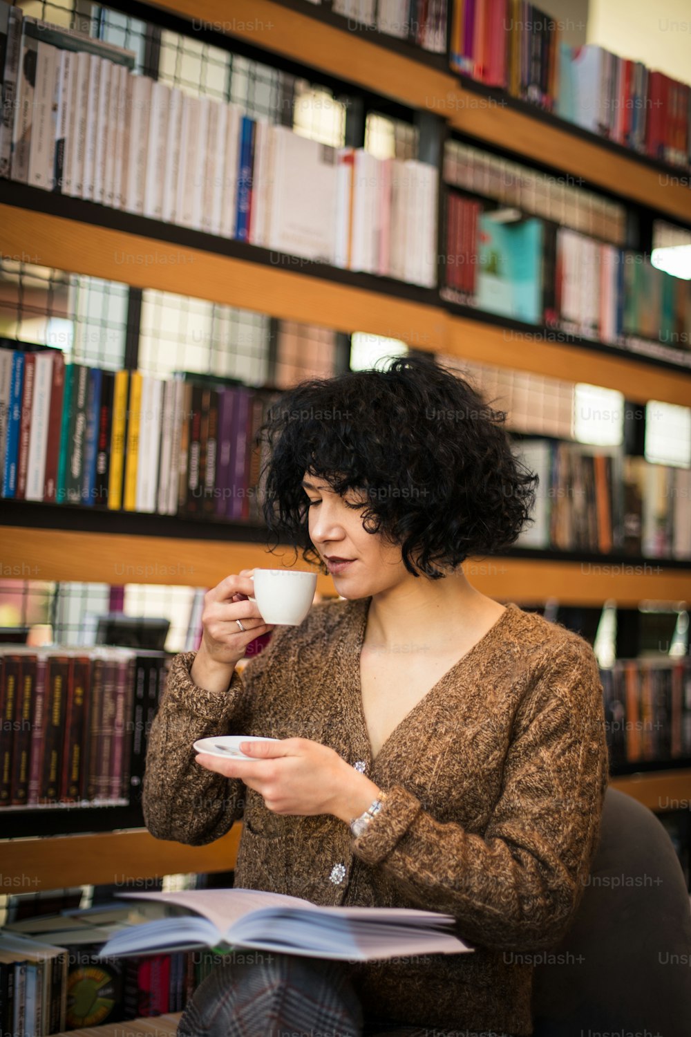 Frau sitzt in der Bibliothek, trinkt Kaffee und liest ein Buch.
