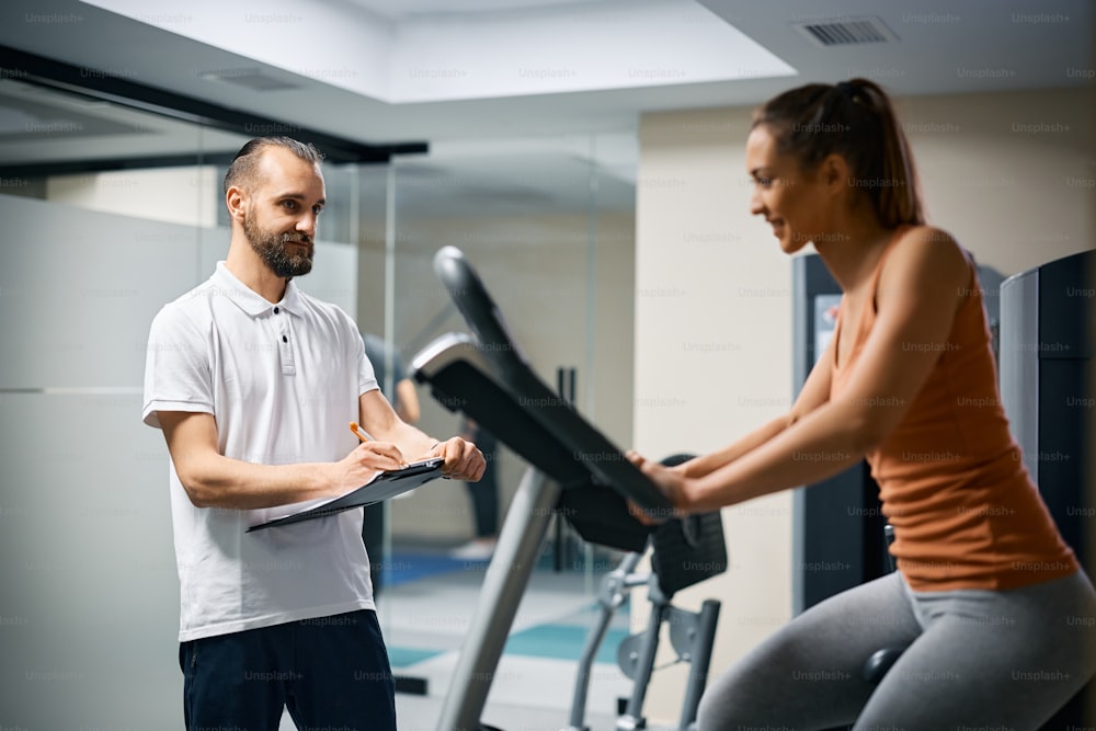 Fitness instructor writing notes while his female client is exercising on stationary bike during sports training at health club.