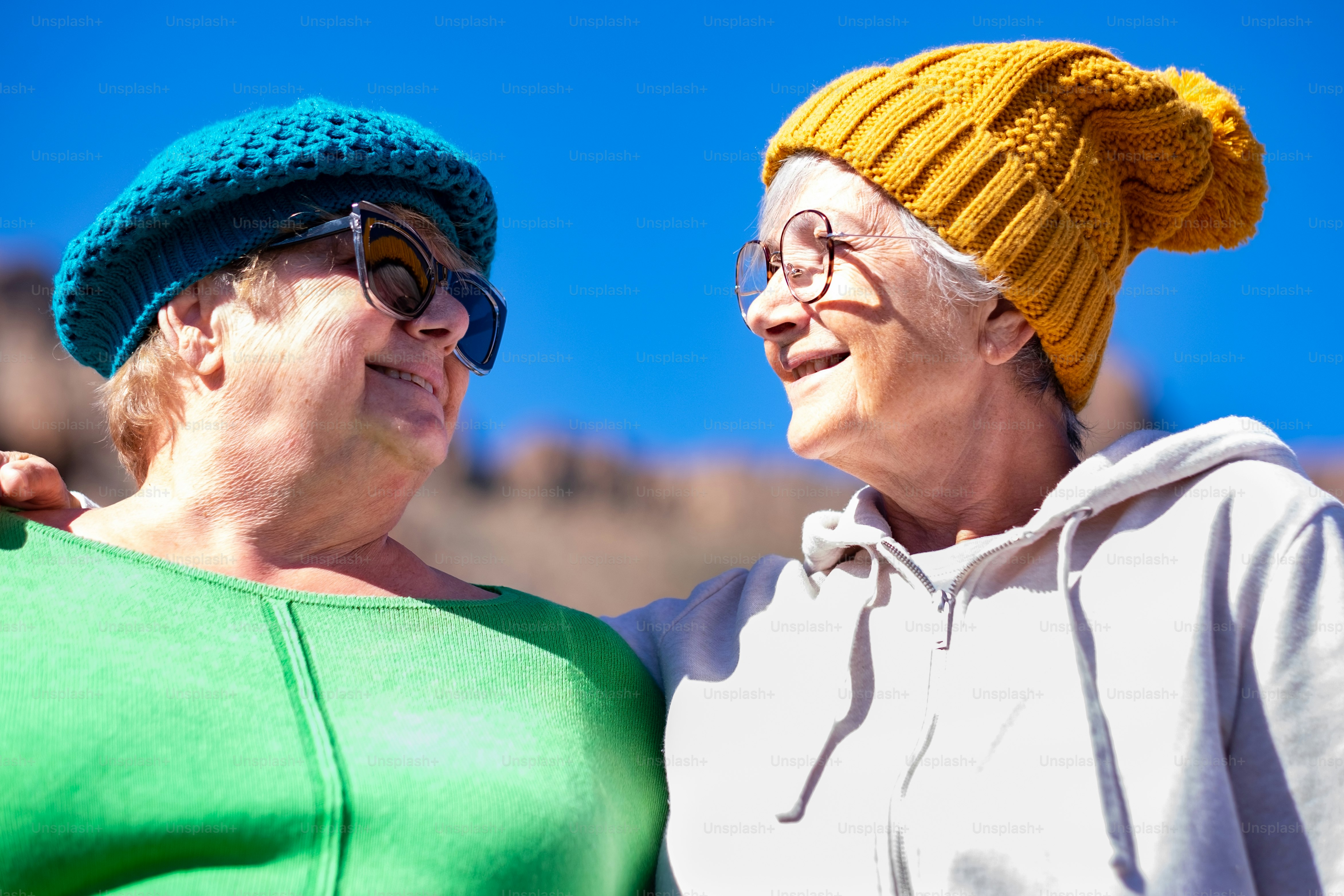 Happy couple of mature elderly women wearing glasses and hats hug looking into the eyes. Smiling carefree senior friends enjoying good time and vacation