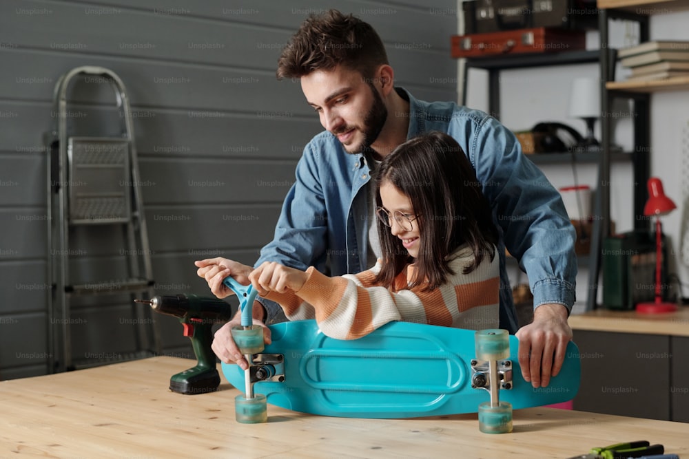 Süßes lächelndes Mädchen mit Handwerkzeug, das Räder des Skateboards auf einem Holztisch in der Garage befestigt, während ein junger Mann im Jeanshemd ihr hilft