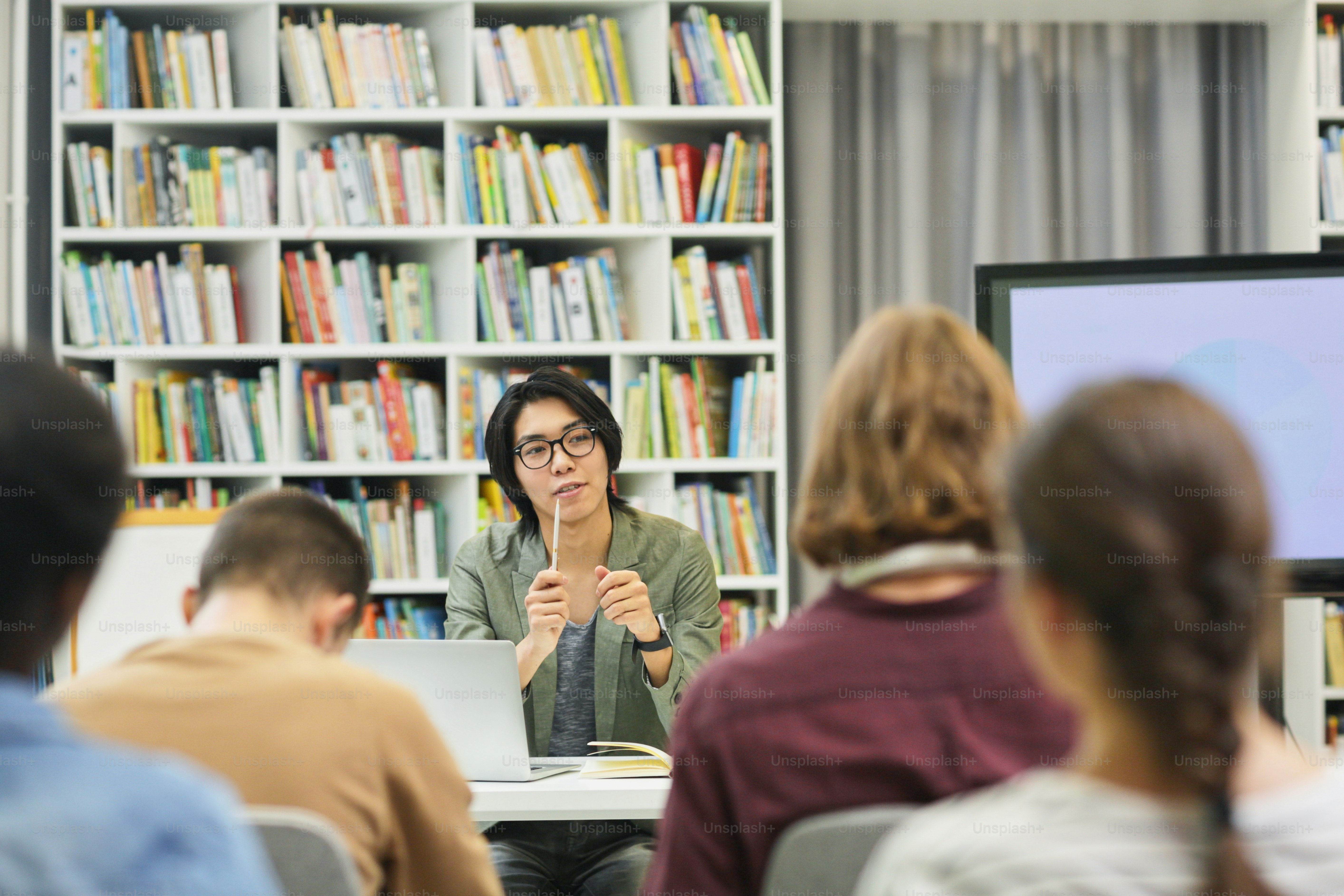 Jeune professeur asiatique à lunettes assis à un bureau et expliquant le nouveau matériel au groupe d’étudiants de la bibliothèque