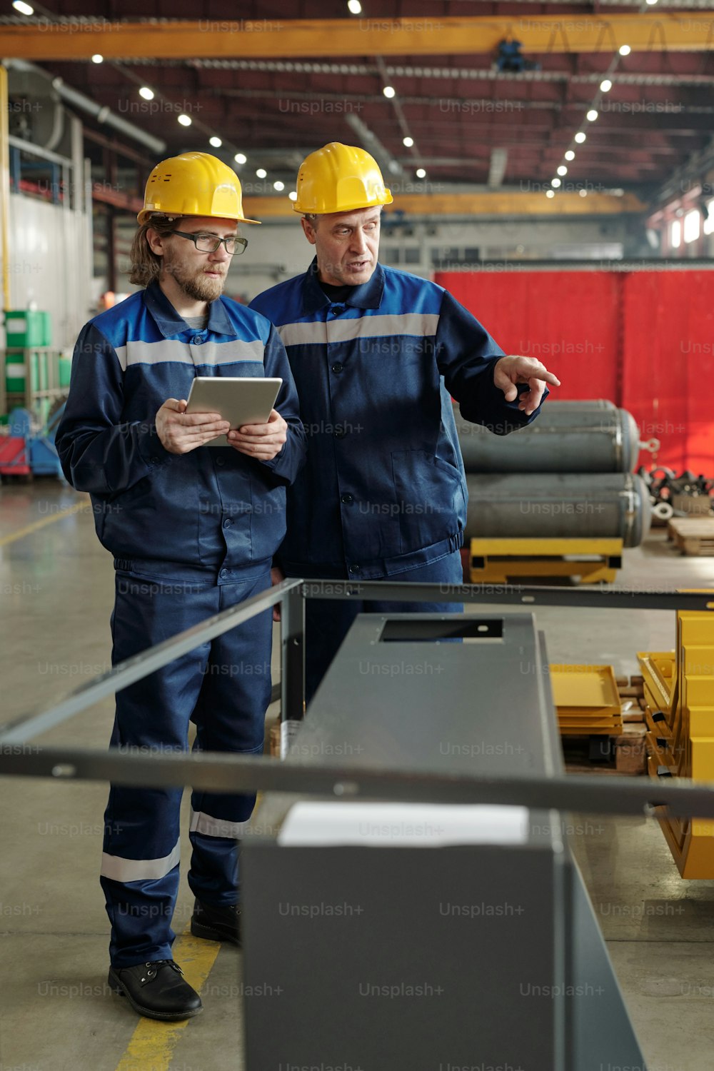Mature foreman in hardhat showing manufacturing equipment to young specialist with tablet while explaining his tasks