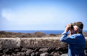 双眼鏡で地平線を眺めている一人の年配の男性の側面図-歩道を屋外で歩く-サボテンのある乾燥した風景-青い海と晴れた日
