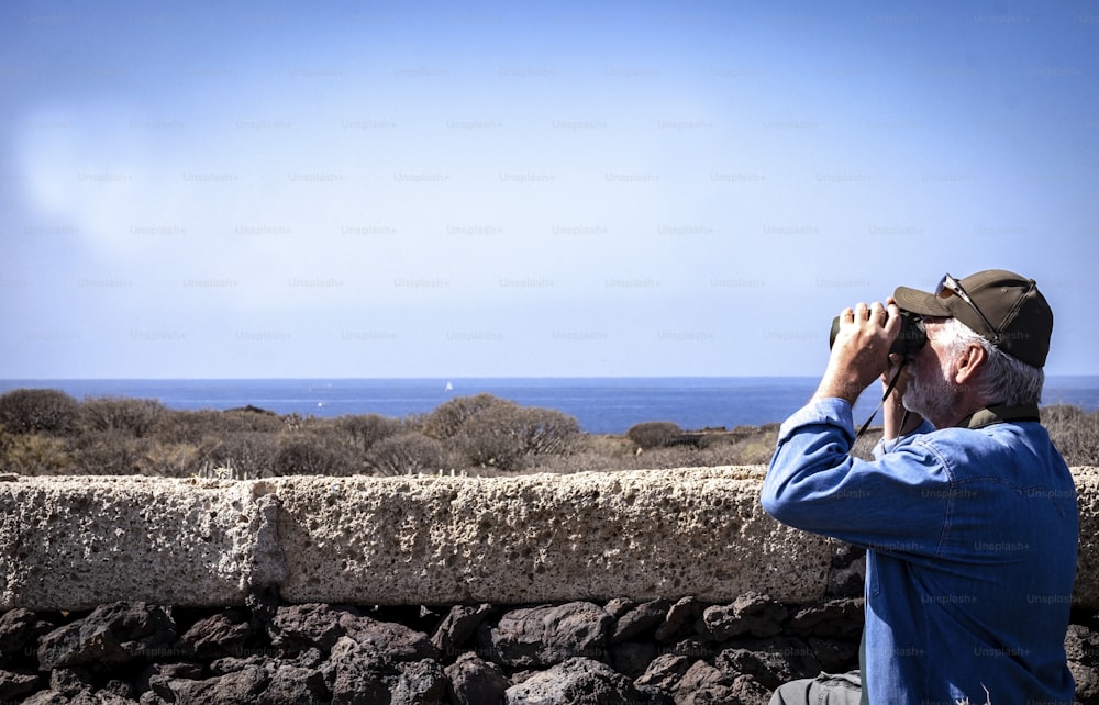 Side view of one senior man with binoculars looking at the horizon - walking outdoor in the footpath - arid landscape with cactus - blue sea and sunny day