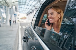 Smiling contented Caucasian lady looking out of taxicab window during phone conversation