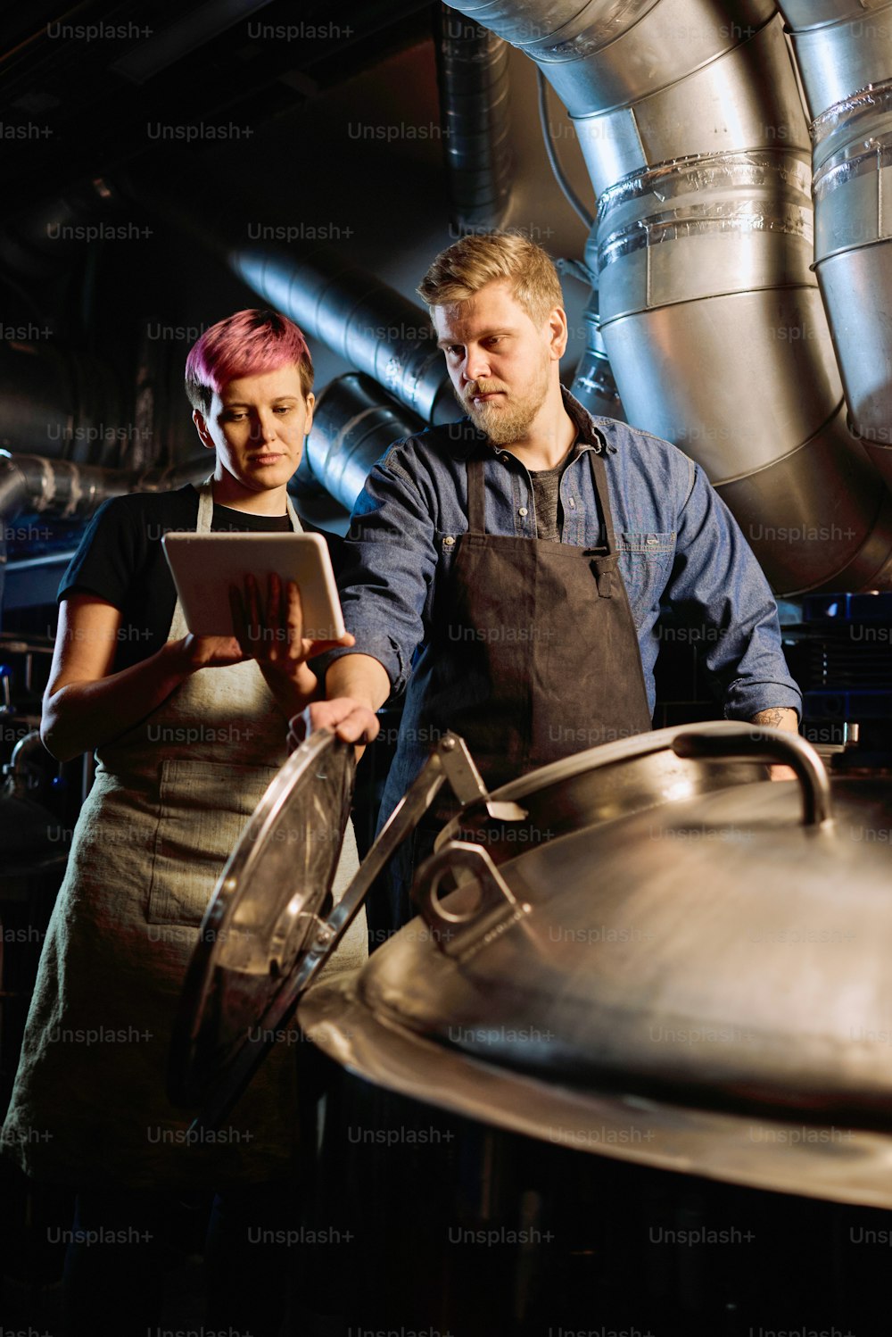 Young woman with tablet scrolling through list of beer processing stages and showing it to her male colleague using special equipment