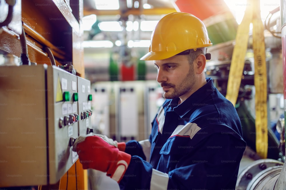 Vista lateral del trabajador caucásico de la planta de energía en ropa de trabajo y con el casco en la cabeza girando el interruptor.