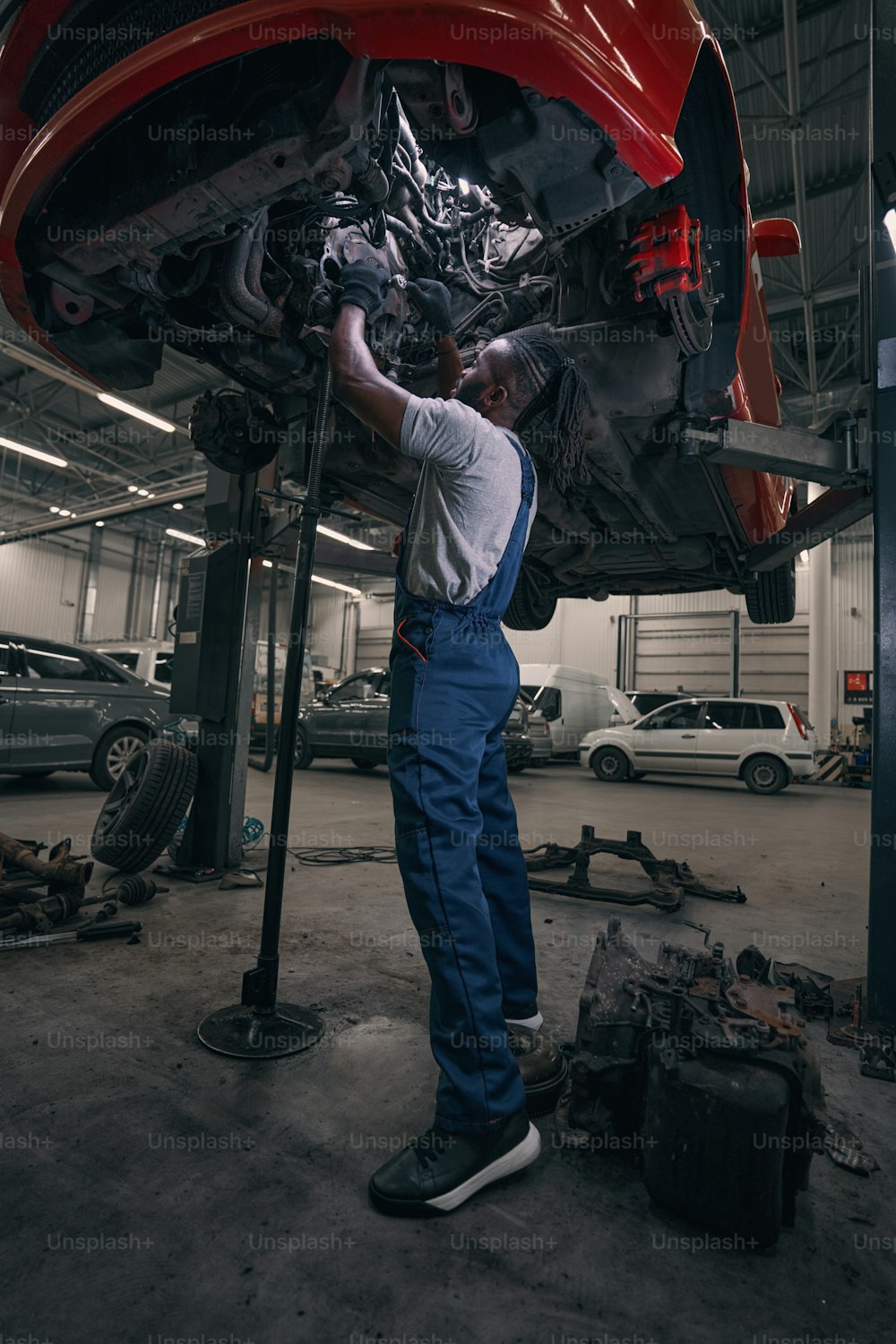 Trabajador afroamericano de pie debajo de un automóvil levantado en un taller de reparación y girando la placa del embrague con una llave inglesa