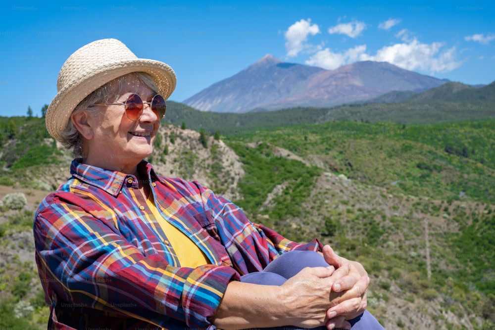 Attractive senior woman with straw hat enjoying outdoor excursion in mountain landscape, smiling. El Teide volcan on background.