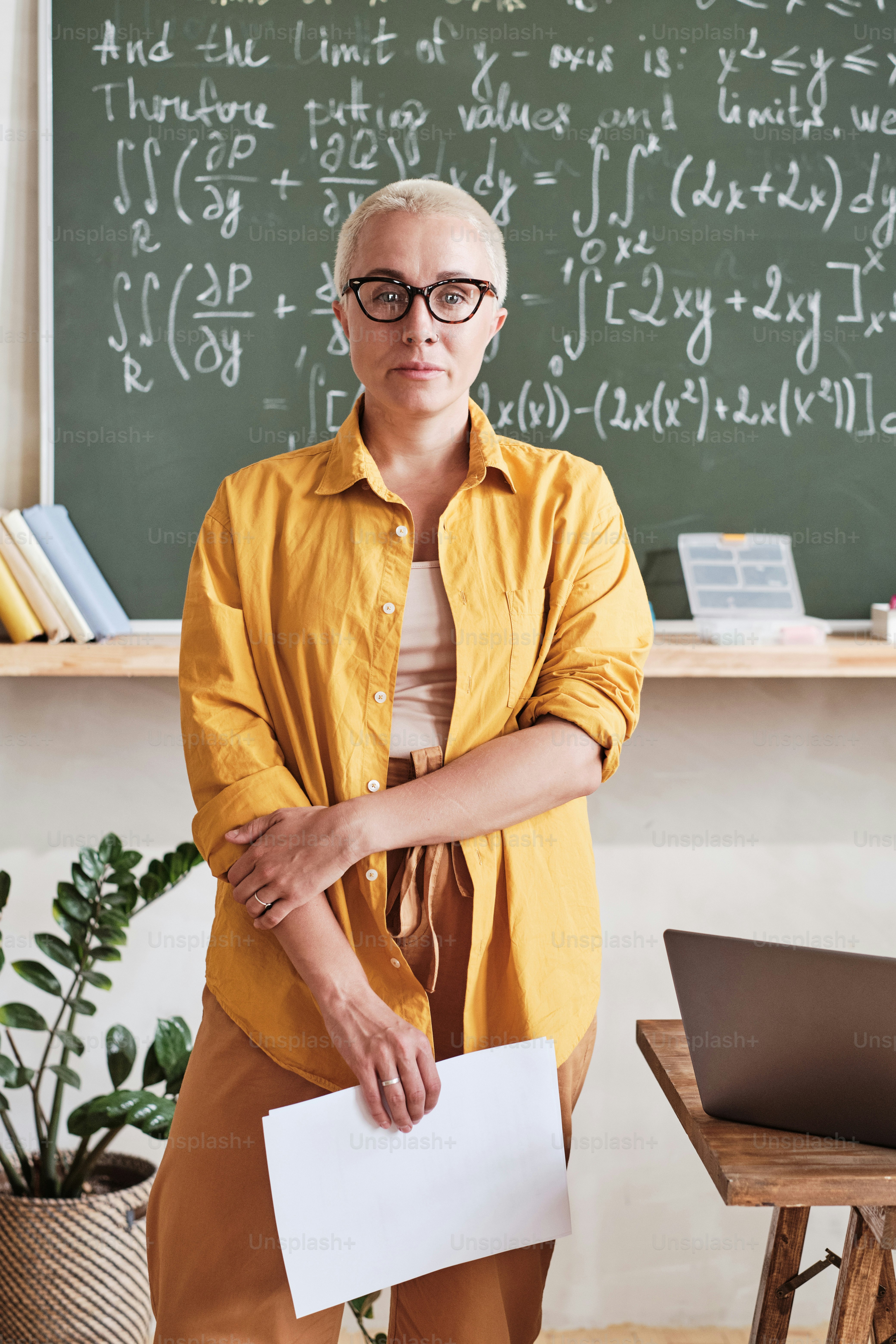 Portrait of confident teacher in eyeglasses standing in the classroom with blackboard in the background