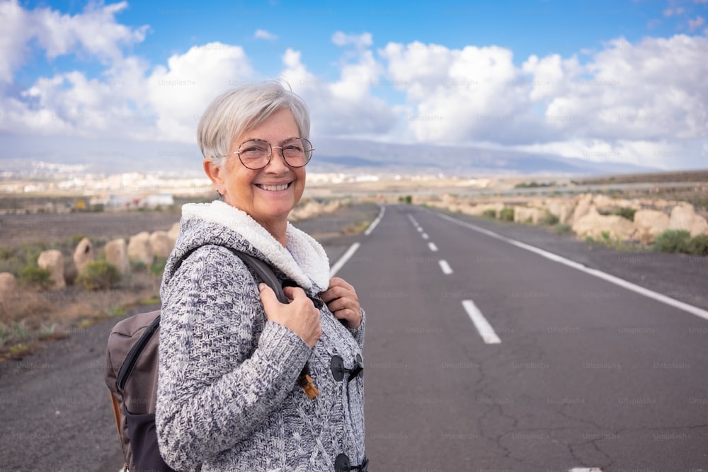 Attractive and happy mature senior woman walking alone in outdoors in country road holding backpack. Elderly smiling woman in empty road with mountain and blue sky in background. Copy space