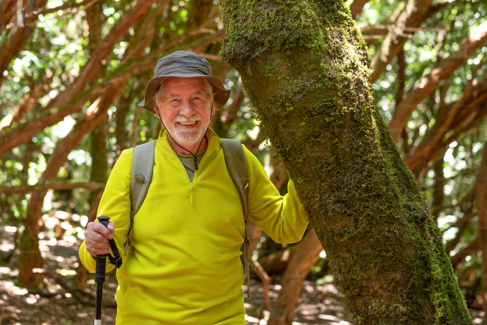 Smiling senior man hiking in the woods touching a moss covered tree trunk enjoying healthy lifestyle - earth day concept. People need to save the planet from deforestation