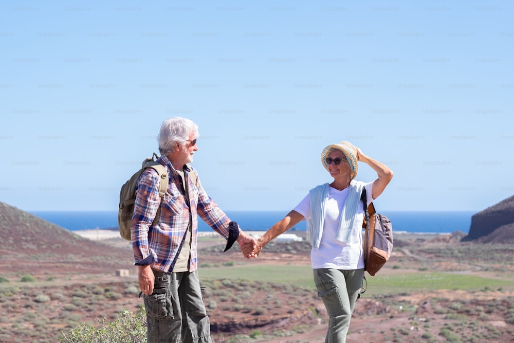 Active smiling senior couple with backpacks in outdoors excursion hiking in mountain looking in the eyes enjoying healthy lifestyle.  Scenic view of sea and mountain background