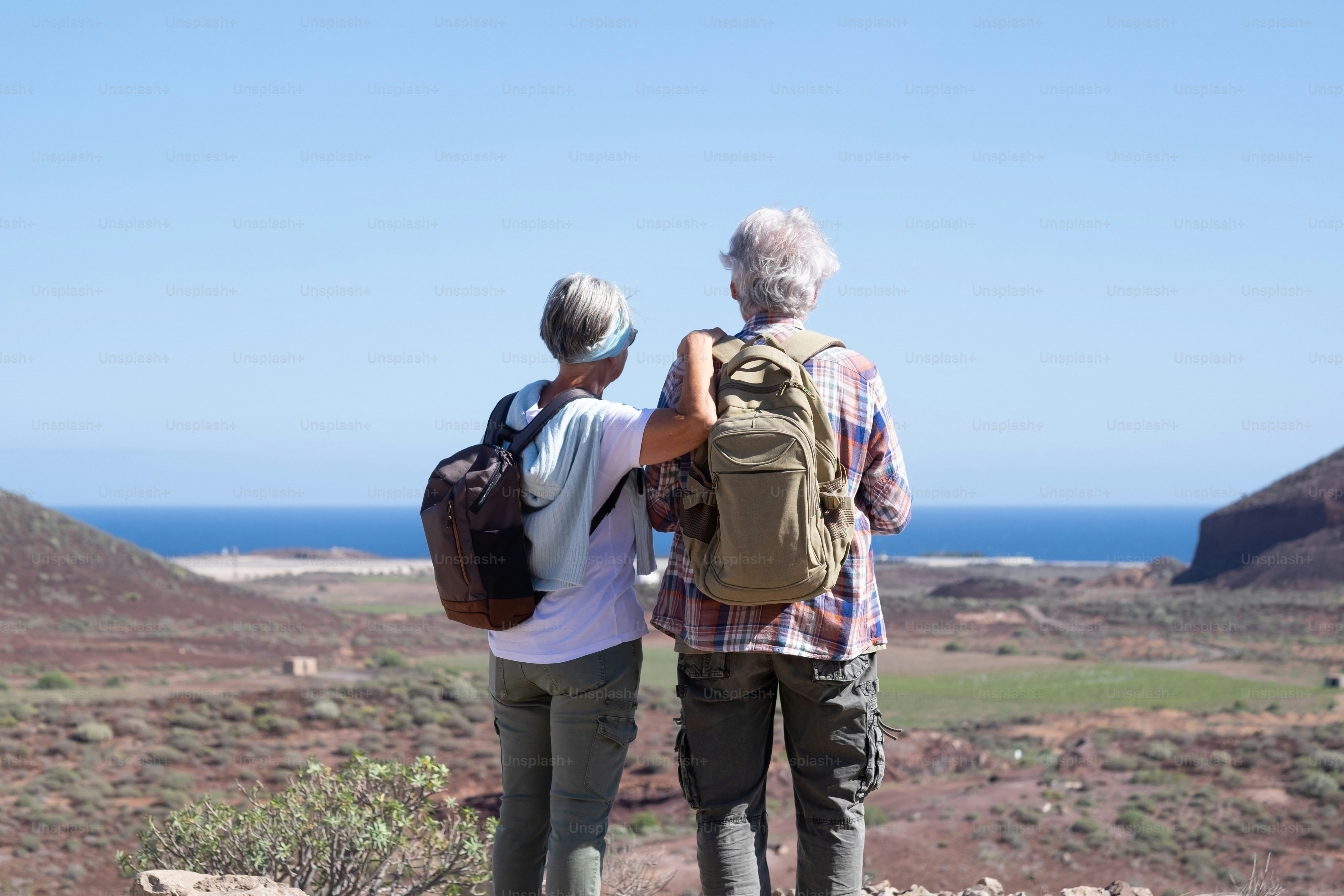 Active senior couple with backpacks in outdoors excursion hiking in mountain looking at horizon enjoying healthy lifestyle.  Scenic view of sea and mountain background
