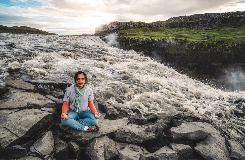 Junge Frau, die Meditationsyoga macht, während sie auf der Seite des Flusses und der großen Wasserfalllandschaft des Dettifoss-Wasserfalls in Island sitzt.