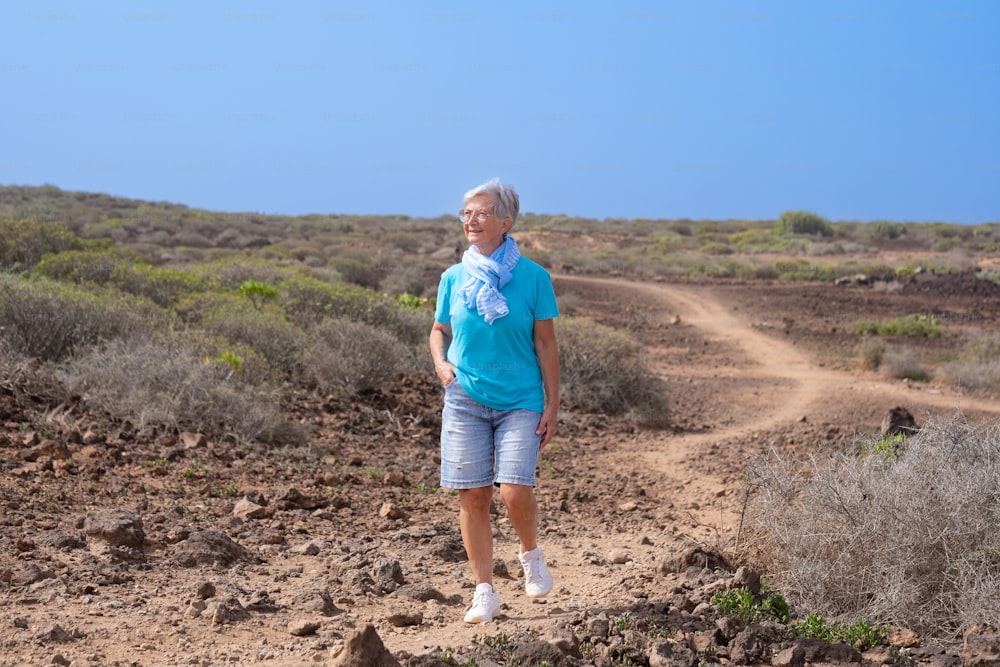 Happy mature senior woman walking outdoor in countryside wearing glasses and blue scarf. Elderly white haired lady enjoying freedom and healthy lifestyle