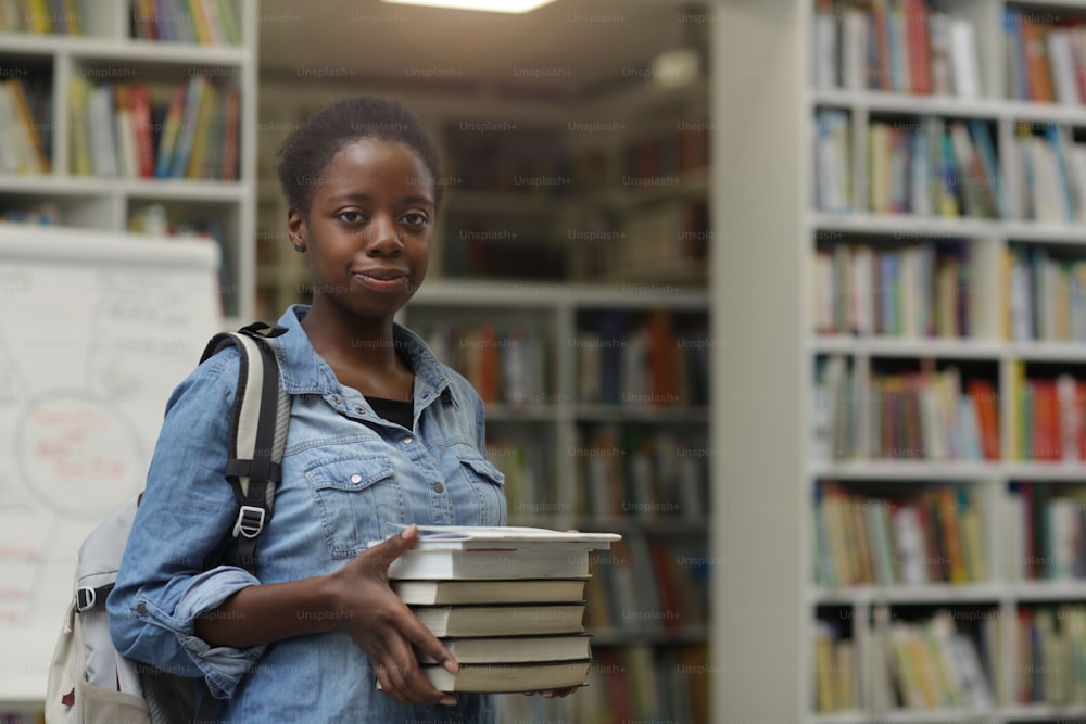 Retrato de una joven africana mirando a la cámara mientras sostiene un montón de libros en sus manos en la biblioteca