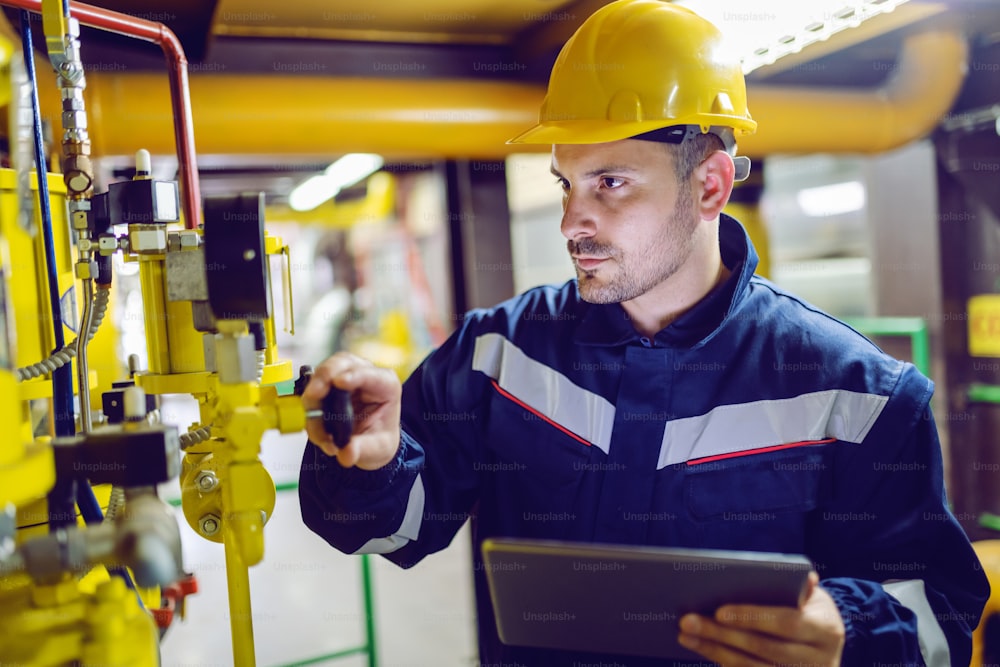 Ernsthafter engagierter Werksarbeiter in Arbeitskleidung und mit Schutzhelm am Kopfschraubventil und Haltetablette.