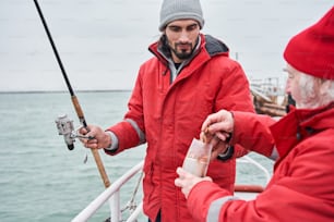 Waist up portrait view of the two colleague men with spinnings standing on the boat and taking out the bait to hook it on the hook. Occupation concept. Stock photo