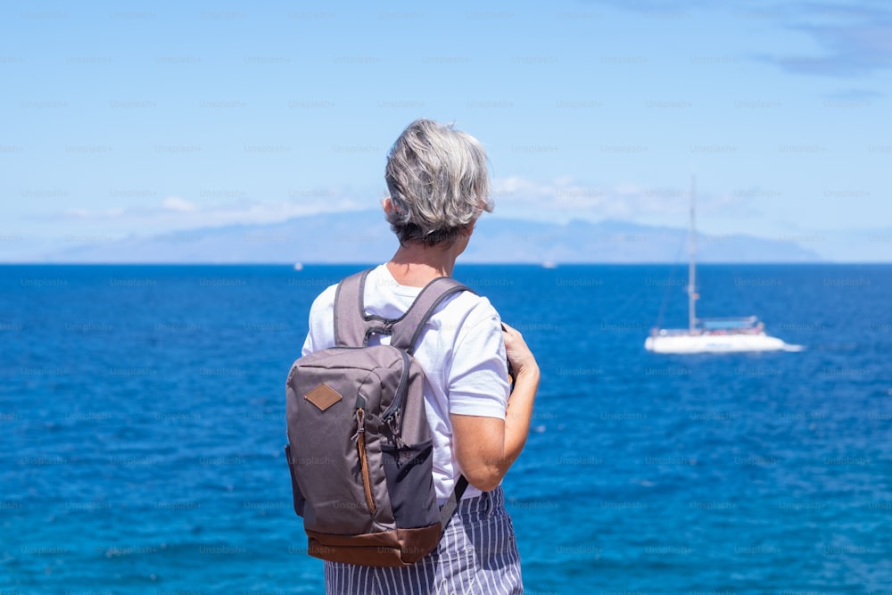 Rear view of senior woman with backpack standing and looking at sea and at the island at the horizon, summer holidays or retirement