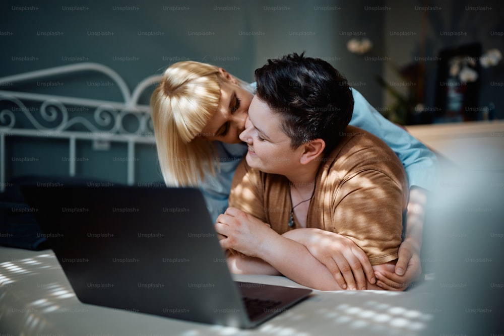 Lesbiana feliz usando computadora portátil mientras su novia la besa en el dormitorio.