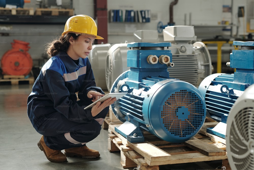 Young serious female engineer in protective helmet and workwear squatting by new industrial equipment and using digital tablet