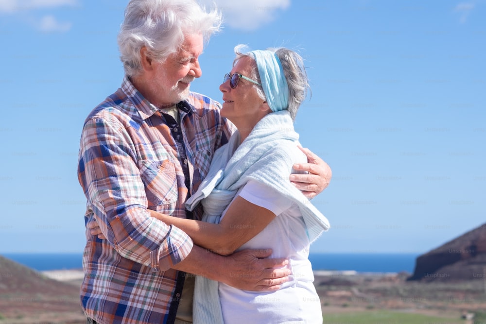Lovely active senior couple hugging in mountain range enjoying healthy lifestyle and sunny day. Two smiling elderly people during vacation, horizon over water.