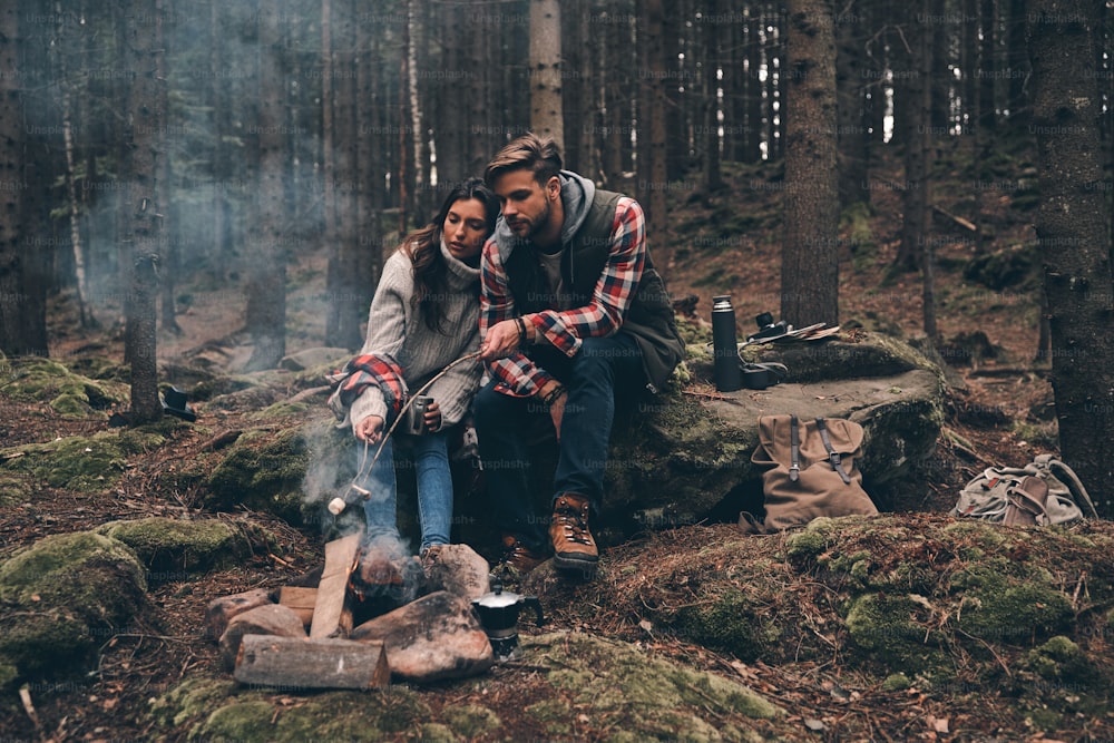 Hermosa pareja joven asando malvaviscos sobre una fogata mientras se sienta en el bosque