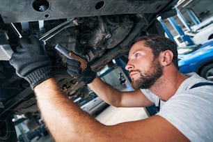 Qualified male technician standing below car and checking its floorpan while shining with flashlight