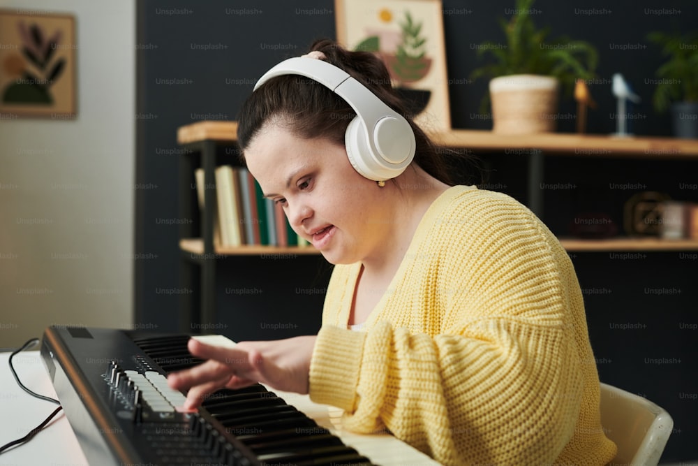 다운 증후군을 앓고 있는 현대 젊은 백인 여성이 전자 키보드의 사운드 설정을 조정하는 캐주얼한 옷과 헤드폰을 착용하고 있다