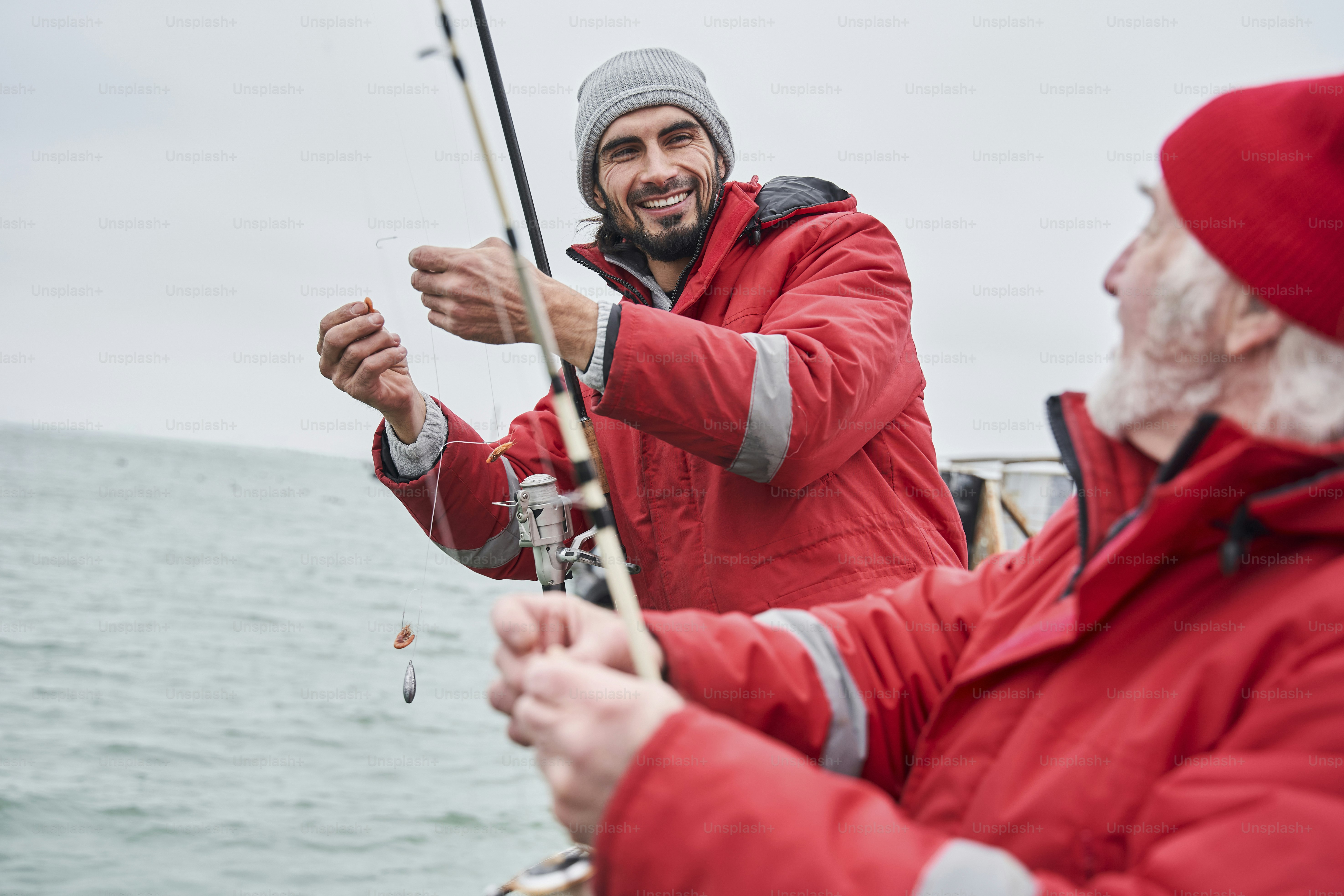 Pesca profesional. Vista de retrato de cintura para arriba del joven sin afeitar diciéndole algo a su colega mayor mientras se prepara para la pesca. Foto de archivo