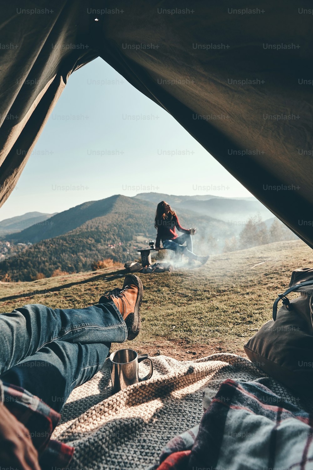 Unkenntlicher junger Mann, der vom Zelt aus den Blick auf die Bergkette genießt, während seine Freundin sich am Lagerfeuer ausruht