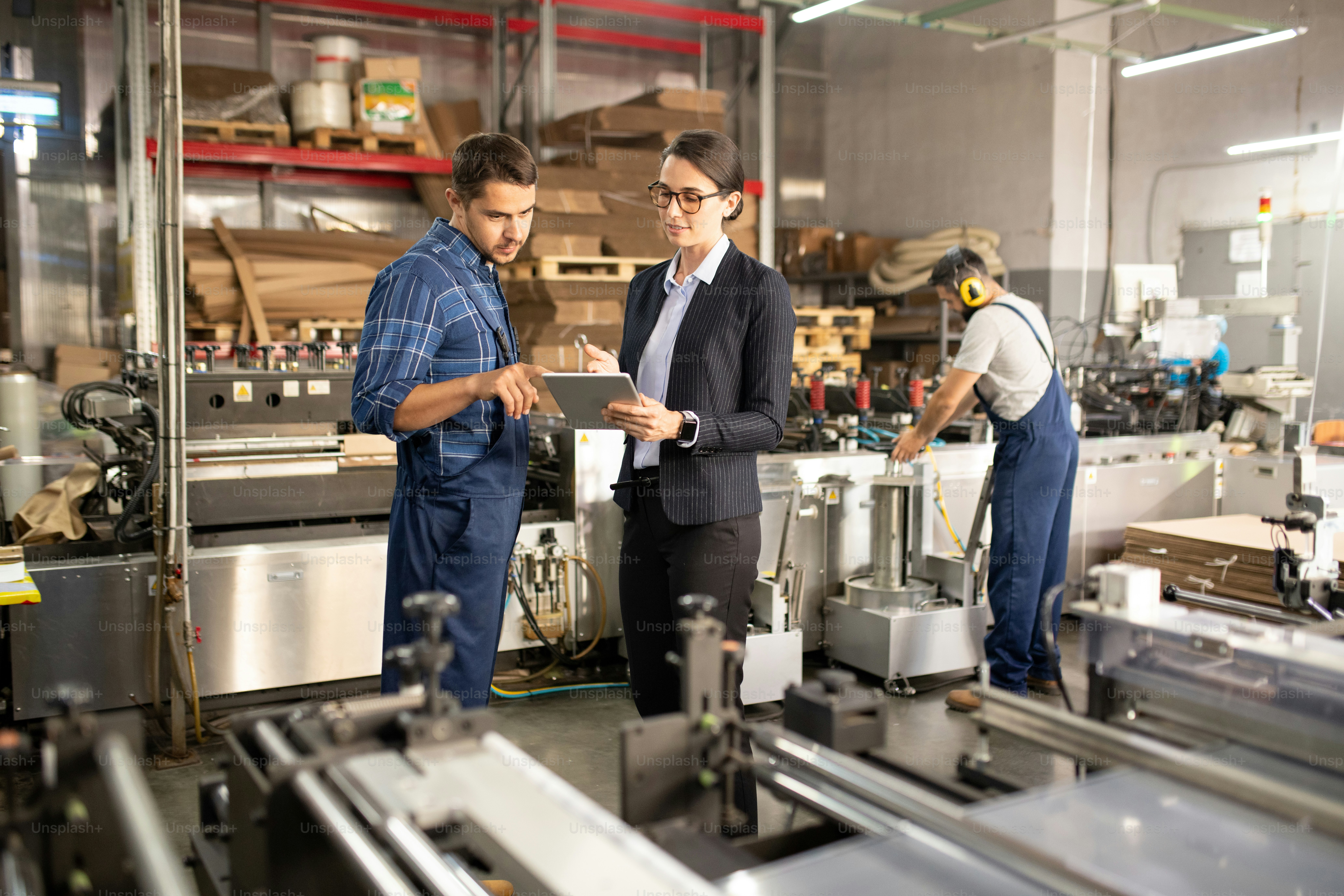 Two young confident workers of processing factory discussing online data in tablet while standing by one of industrial machines