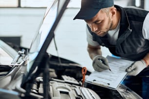 Jeune mécanicien vérifiant l’huile de voiture et prenant des notes tout en travaillant dans un atelier de réparation automobile.
