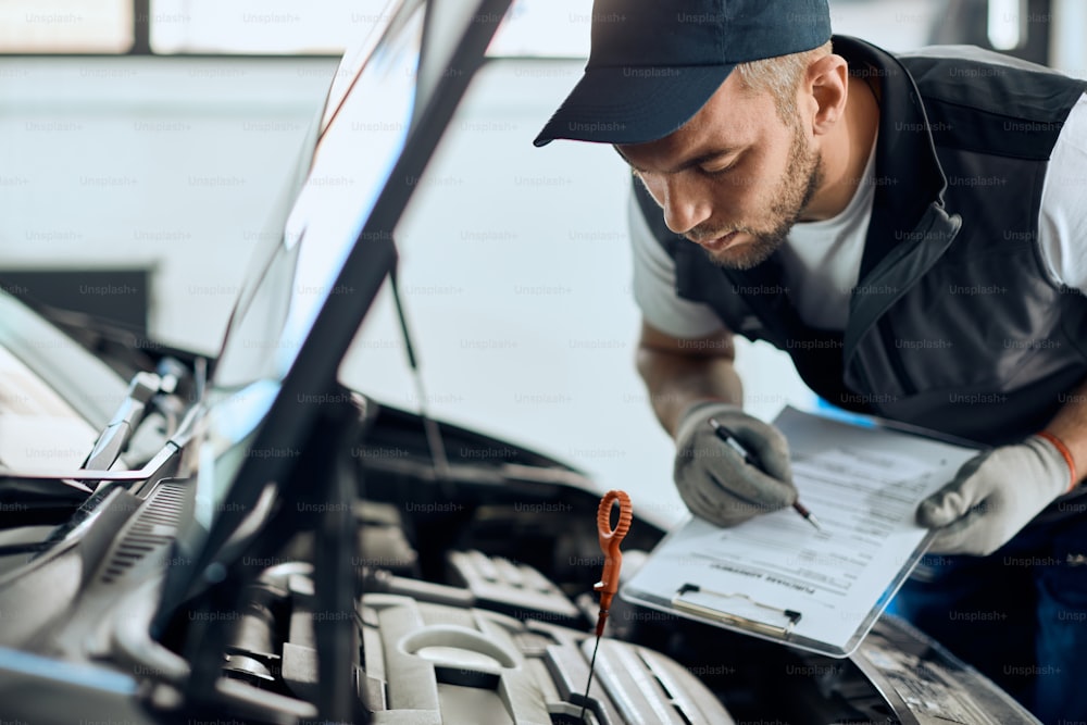 Giovane meccanico che controlla l'olio dell'auto e prende appunti mentre lavora in un'officina di riparazione auto.