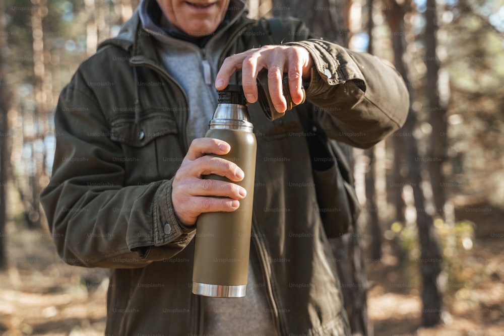 Close up view of the cheerful senior man preparing to pouring warm tea from the thermos with kind smile. Male is blurred, focus on the bottle