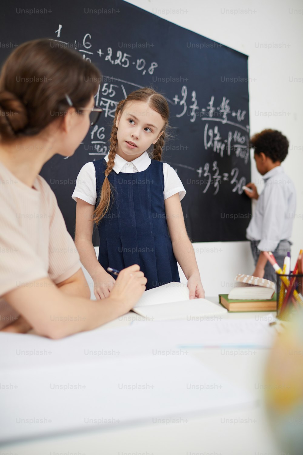 Young teacher sitting at the table and talking to schoolgirl while boy working at the blackboard in the background