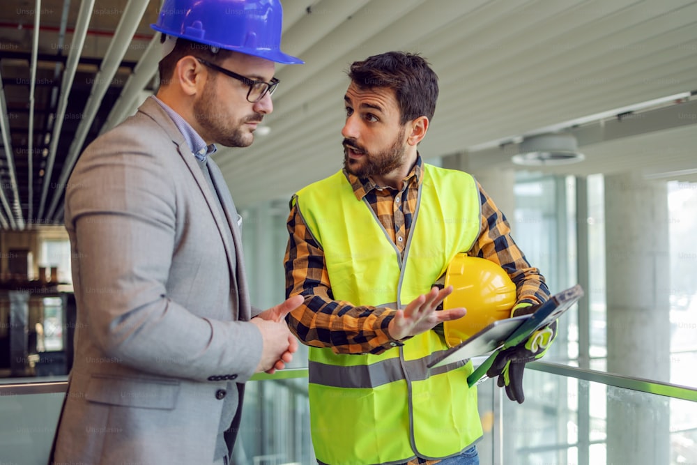 Construction worker holding tablet and explaining his supervisor how works are going. Building in construction process interior.