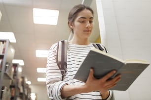 Serious student with backpack behind her back standing and reading a book in the library