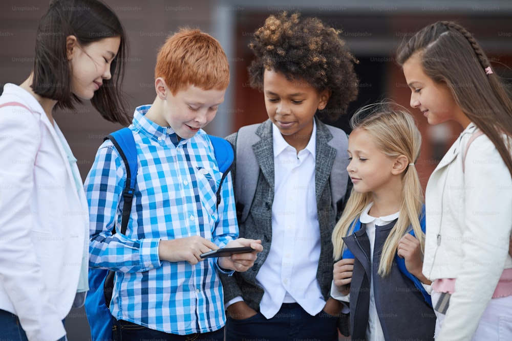 Grupo de niños de la escuela multiétnica que juegan juegos en línea en el teléfono móvil mientras están de pie al aire libre