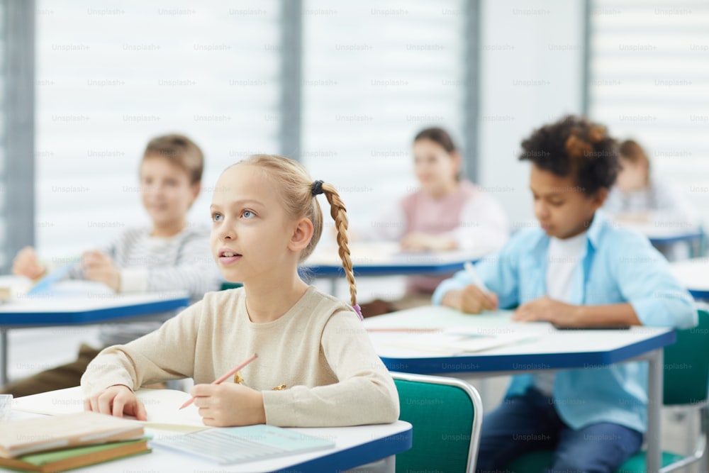 Menina curiosa com cabelos loiros sentados na mesa da escola ouvindo seu professor, retrato horizontal, espaço de cópia