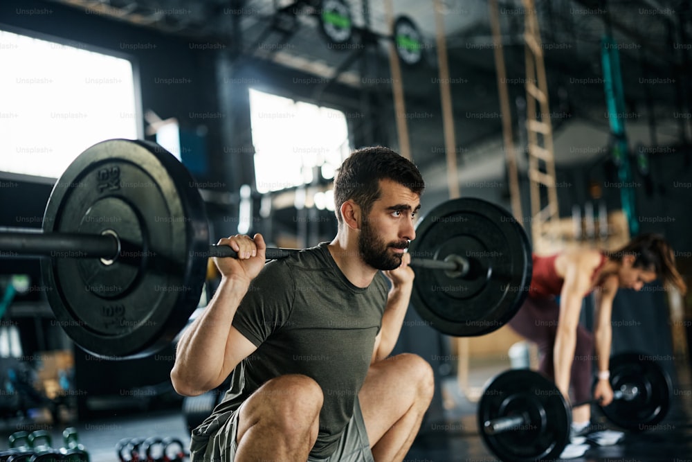 Athletic man using barbell while practicing squats during cross training in a gym.